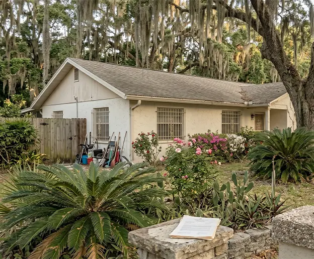 Single-story Florida house with yard tools leaning against the wall and probate documents sitting on a front pillar